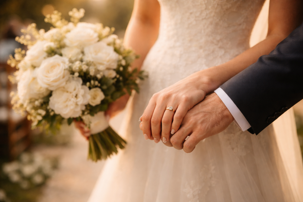 mãos do casal entrelaçadas durante cerimônia de casamento com buquê ao fundo desfocado